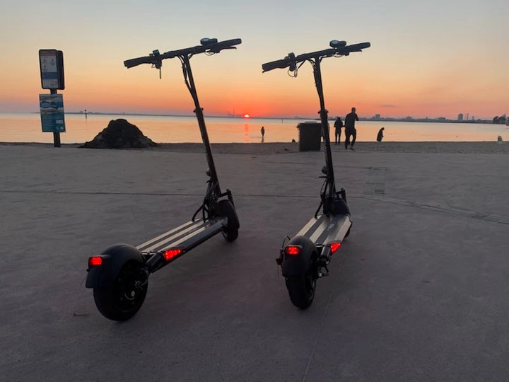 Two black EMOVE Cruiser electric scooters parked on the St Kilda boardwalk at sunset, highlighting their sleek design and versatility for scenic rides. Customer photo by Dave Thomas.
