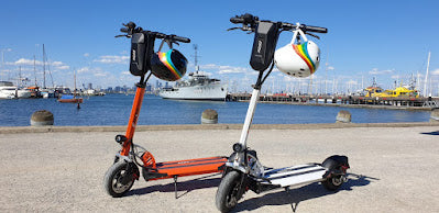 Orange and white EMOVE Cruiser E-Scooters parked near the waterfront at Williamstown, featuring black and white rainbow helmets hanging on the handlebars. Customer photo by Sharon Maguire.