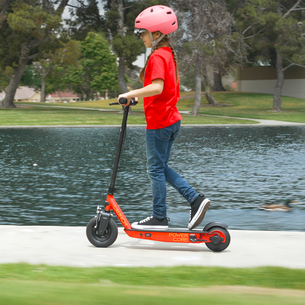 Young girl riding a Razor Launch electric scooter in a park, wearing a pink helmet, gliding smoothly along a path beside a scenic lake.