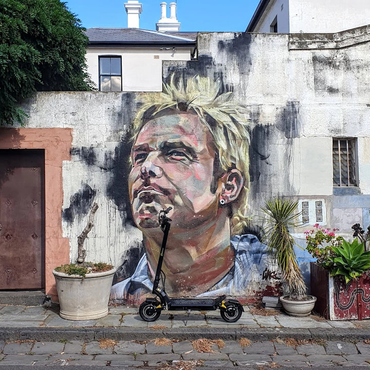 Bexly Blackhawk electric scooter parked in front of a mural of the late cricket legend Shane Warne, located in a St Kilda laneway.
