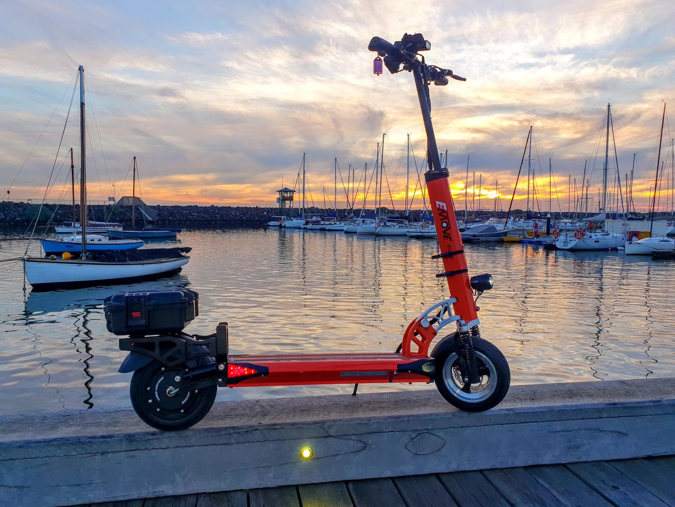 Orange EMOVE Cruiser electric scooter with storage box and accessories parked on St Kilda Pier, overlooking the penguin colony at sunset, showcasing versatile urban mobility and scenic views.