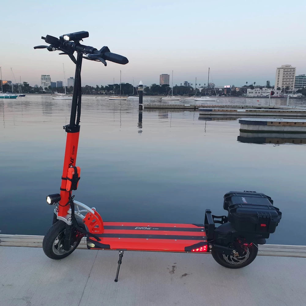 Orange EMOVE Cruiser electric scooter with storage case attached, displayed by the St Kilda waterfront, demonstrating long-range delivery capability.