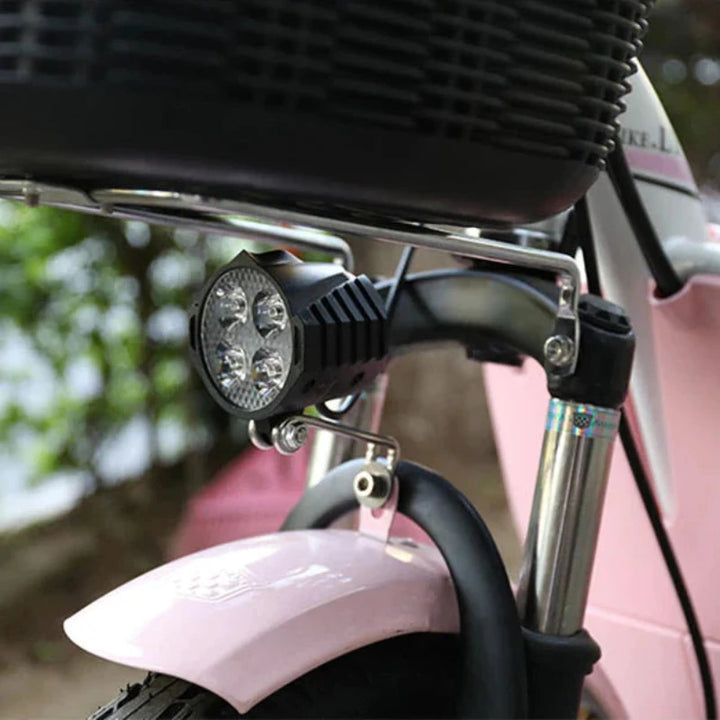 E-bike headlight with horn fitted to a front fork of a pink electric bicycle. The compact 12W light provides enhanced brightness with a sturdy mounting system for secure attachment.