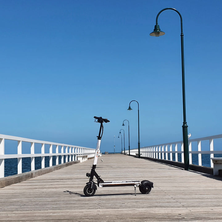 White EMOVE Touring electric scooter parked on Kerferd Road Pier in Albert Park, Melbourne, under clear, sunny skies with vibrant blue hues overlooking Port Phillip Bay on a beautiful day.