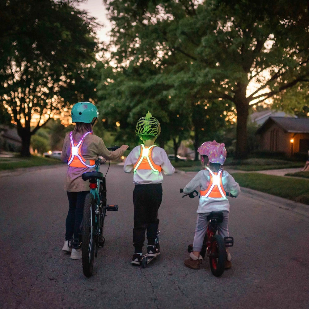 Three children riding bikes and scooters wearing the TracerJR LED safety vests, glowing in vibrant colors at dusk for maximum visibility and safety.