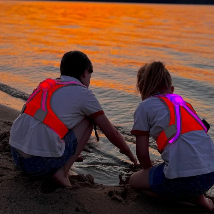 Two children playing by the water's edge, wearing TracerJR LED safety vests in pink and purple, providing visibility during low-light conditions.