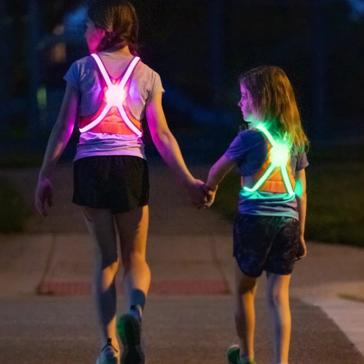 Two children holding hands while crossing the road at dusk, wearing TracerJR LED safety vests in bright multicolour LED modes for maximum visibility.