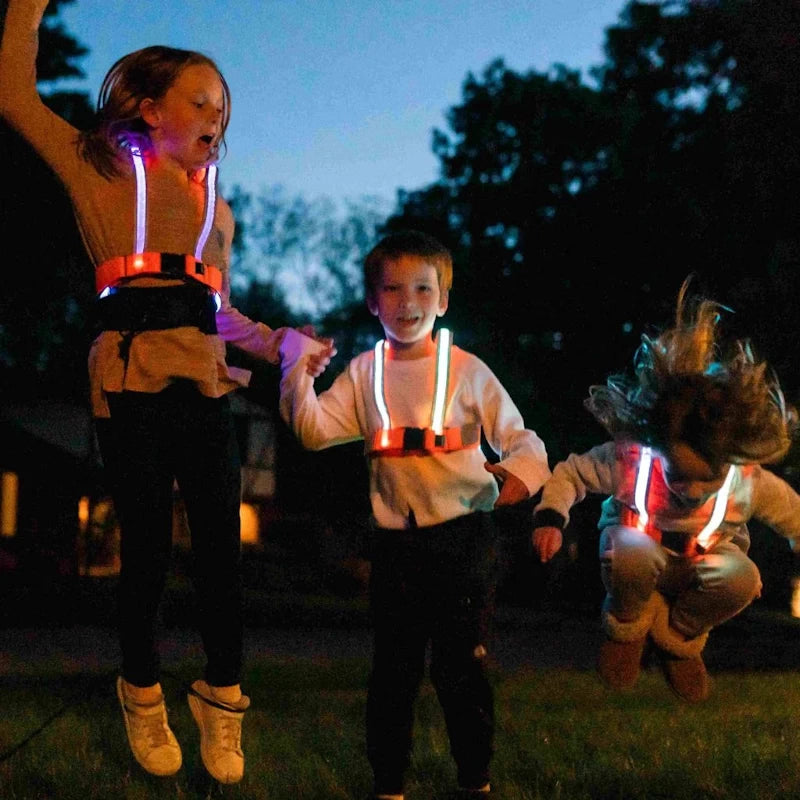 Three children playing and jumping on the grass at dusk, wearing the TracerJR LED safety vests with reflective orange panels and glowing lights.