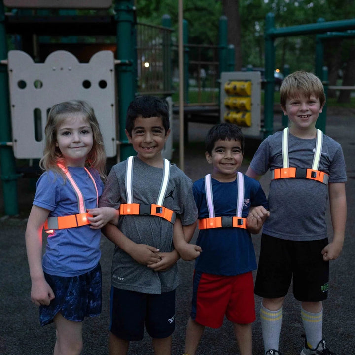 Four kids standing together at a playground at dusk, wearing the TracerJR LED safety vests with bright white and pink LEDs for visibility.