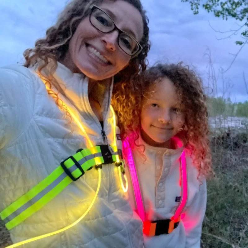 A mother and daughter wearing matching TracerJR LED safety vests, glowing in yellow and pink, enjoying an evening walk together