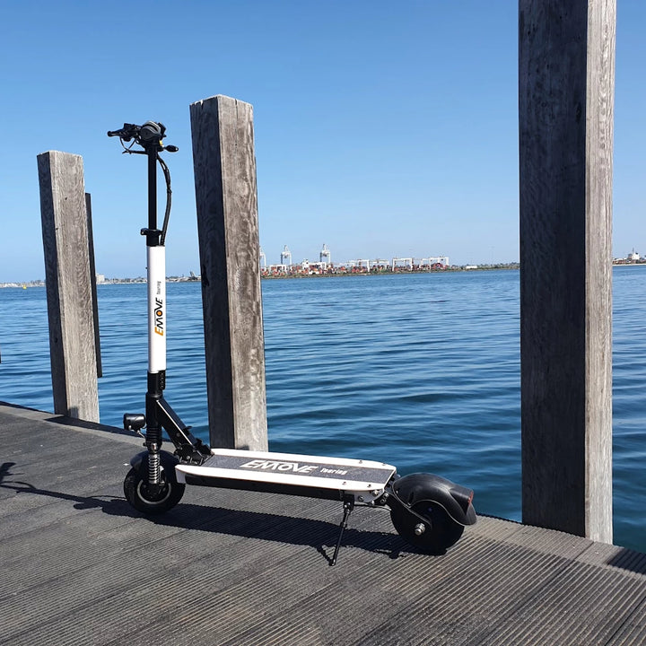 White EMOVE Touring electric scooter parked on the landing stage of a Port Melbourne pier, overlooking the calm waters of Port Phillip Bay with a cloudless blue sky backdrop.